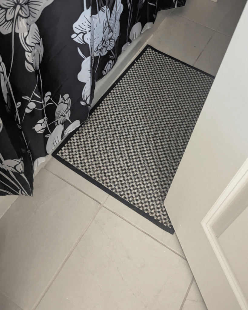 Black and white houndstooth woven bath mat on tile floor in front of a floral shower curtain