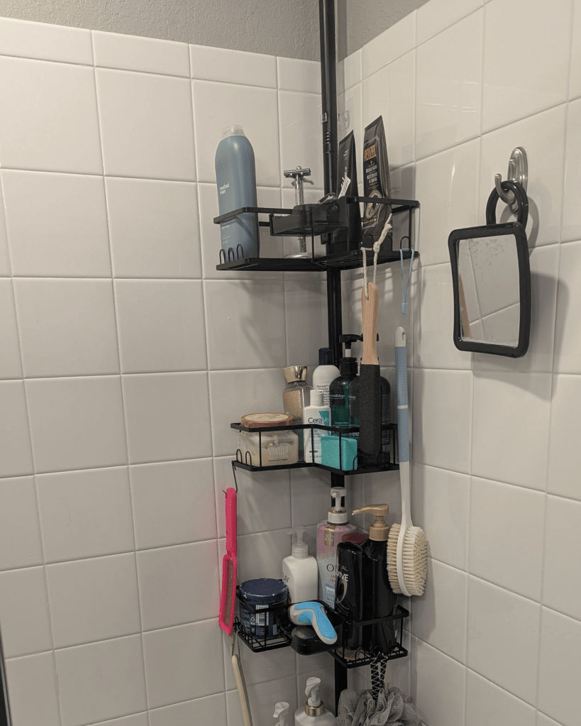 Black tension pole shower caddy with multiple shelves organizing shampoo and bath products in a shared shower